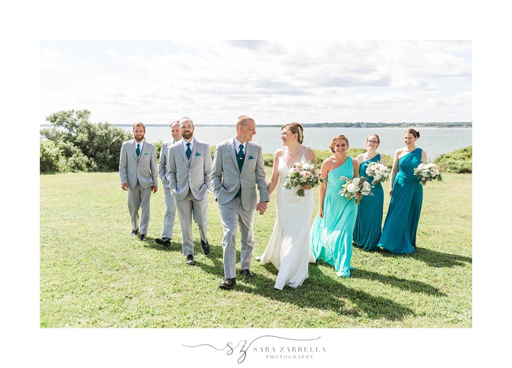 bridal party in teal gowns walks with bride and groom at Brenton Point photographed by Sara Zarrella Photography