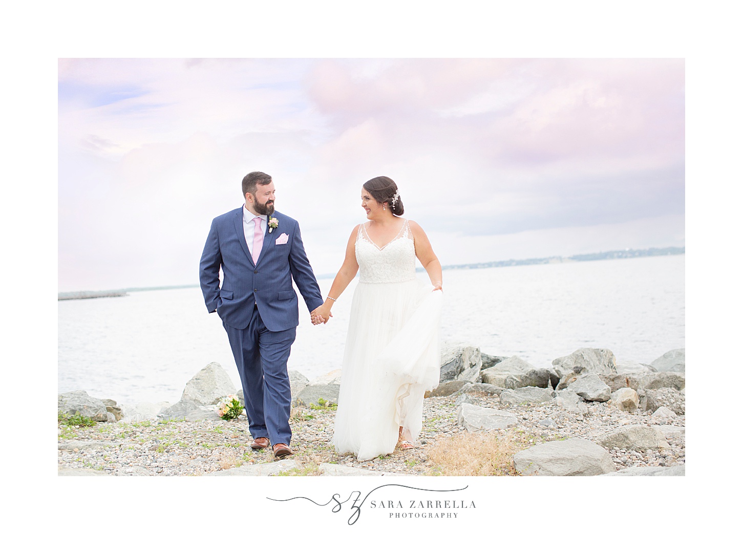 bride and groom walk along rocks by Narragansett Bay