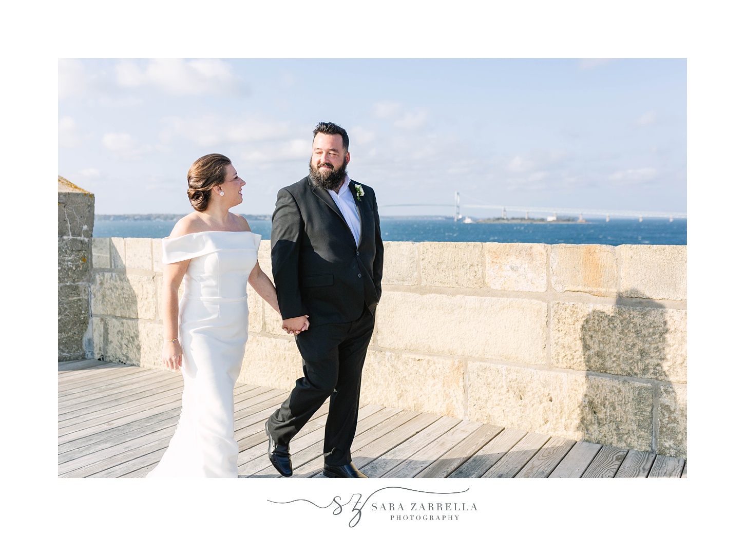 newlyweds walk along top of Fort Adams with bay behind them