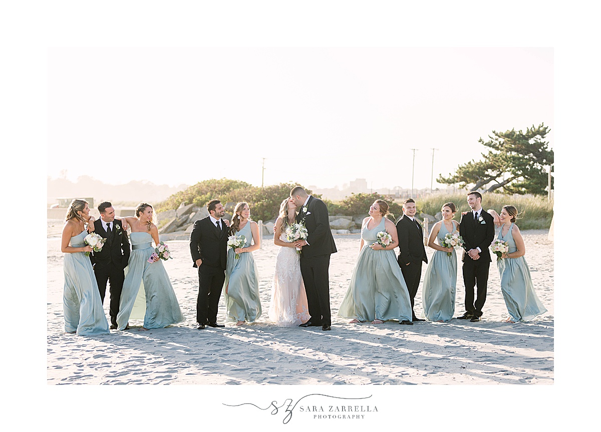 wedding party walks on beach while newlyweds kiss