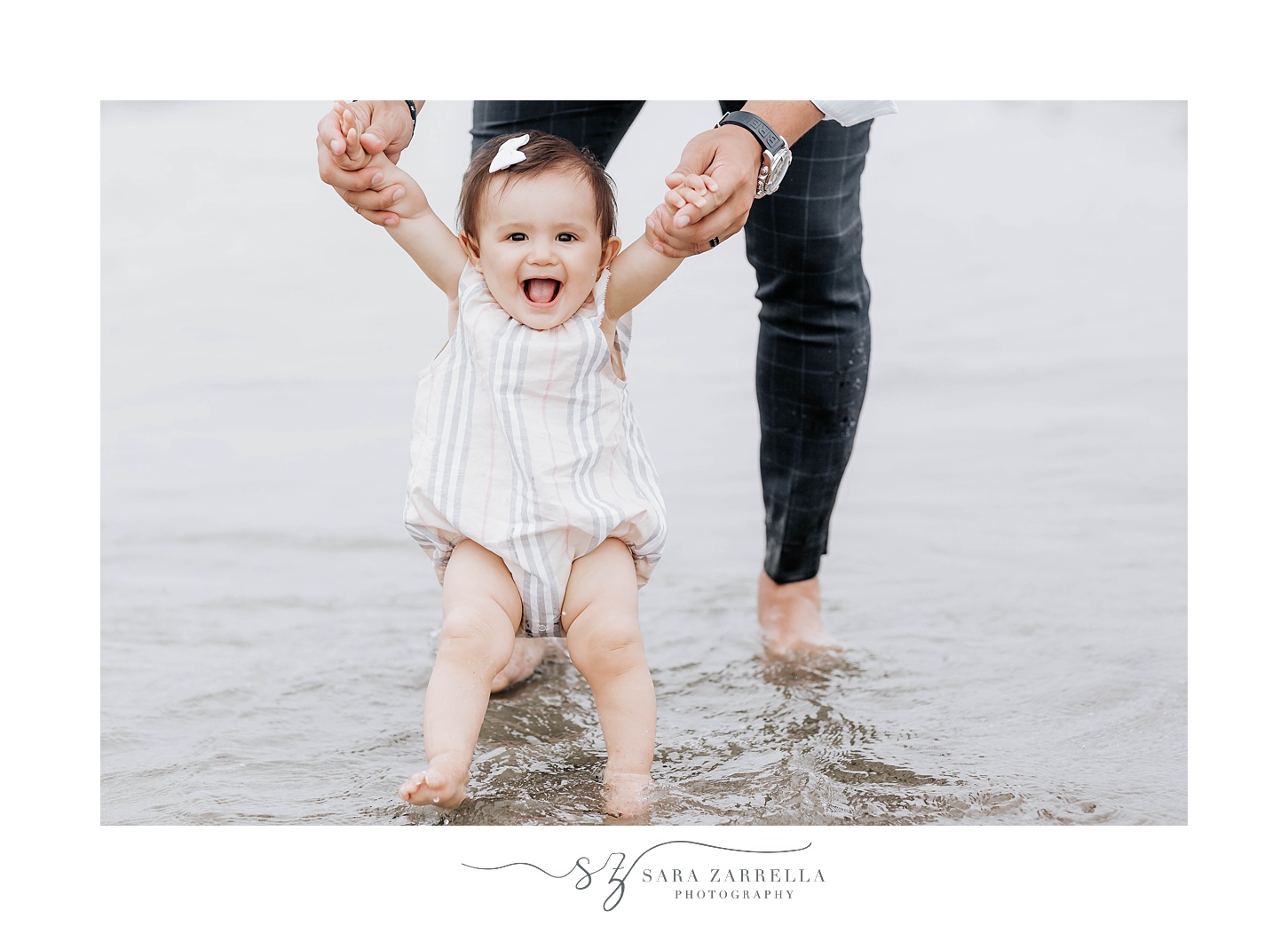 baby stands up on beach with parent's help during beach minis with Sara Zarrella Photography