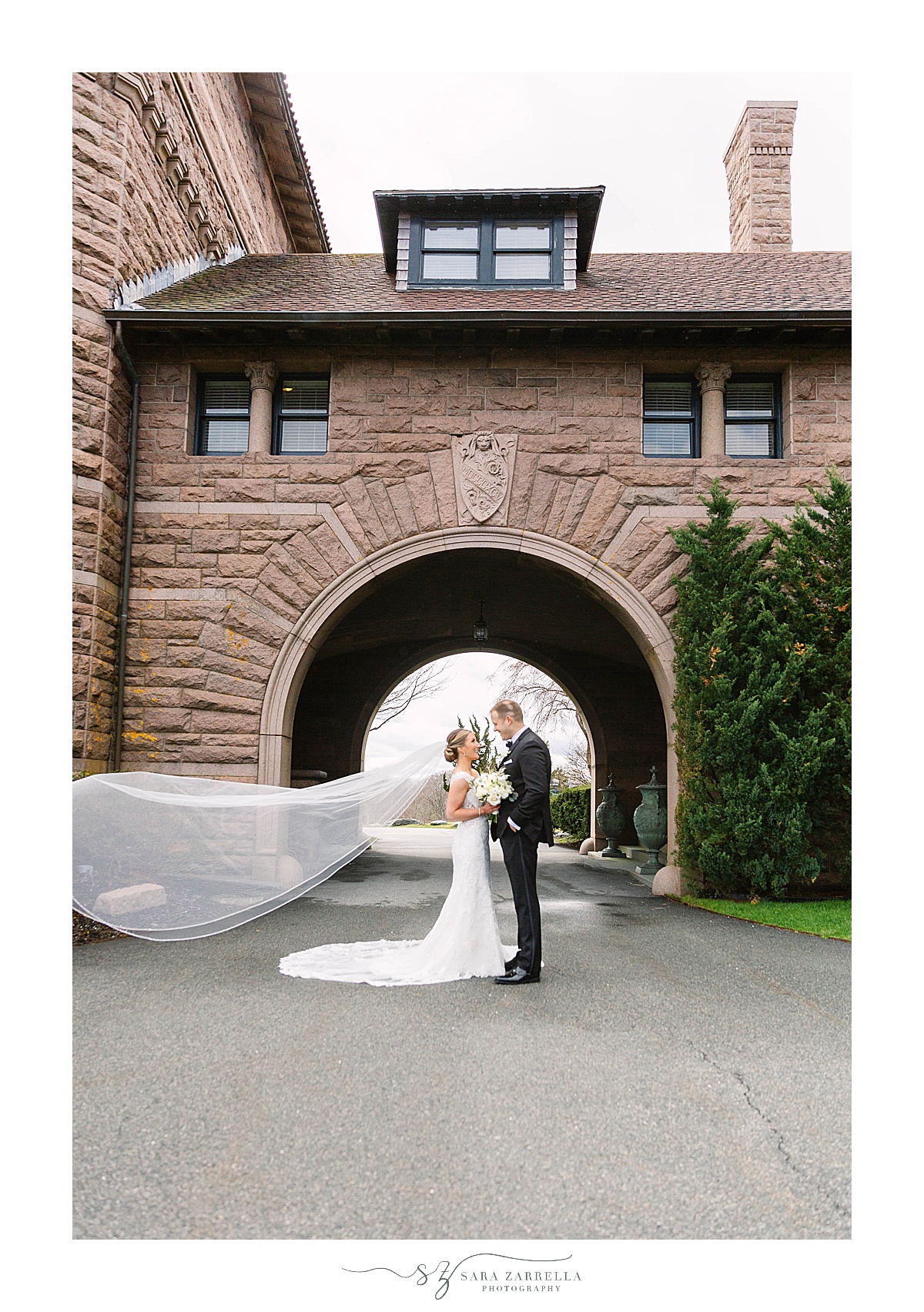 bride and groom pose outside RI venue with bride's veil floating