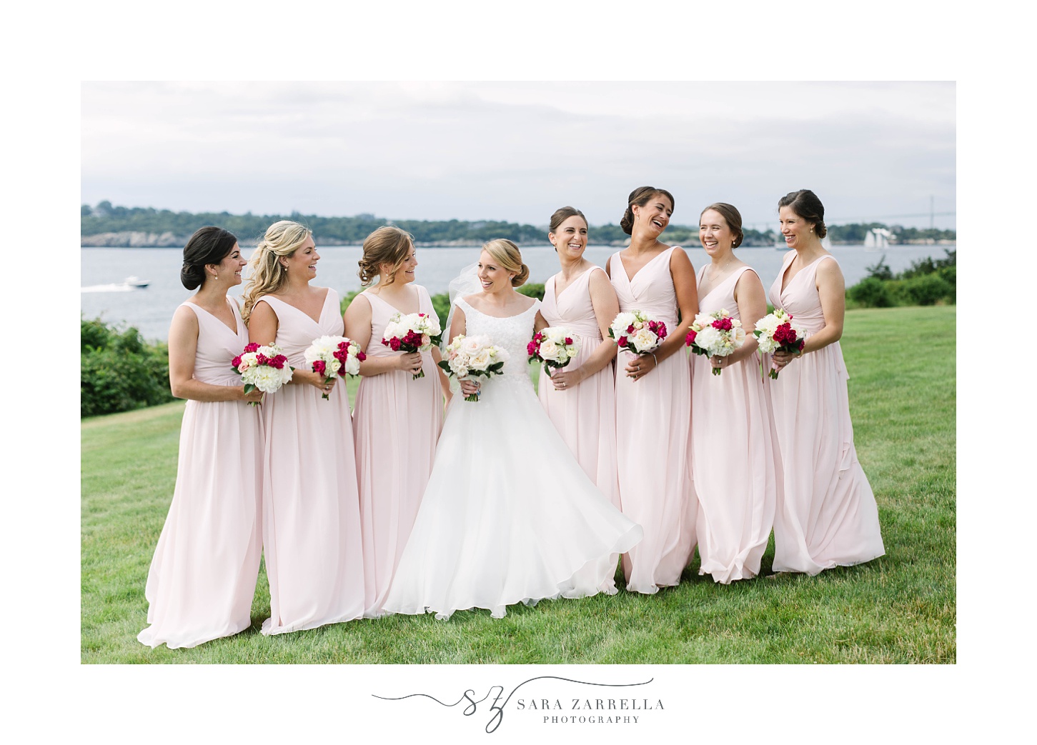 bride and bridesmaids in pink gowns hold bouquets with red flowers along waterfront of OceanCliff Hotel