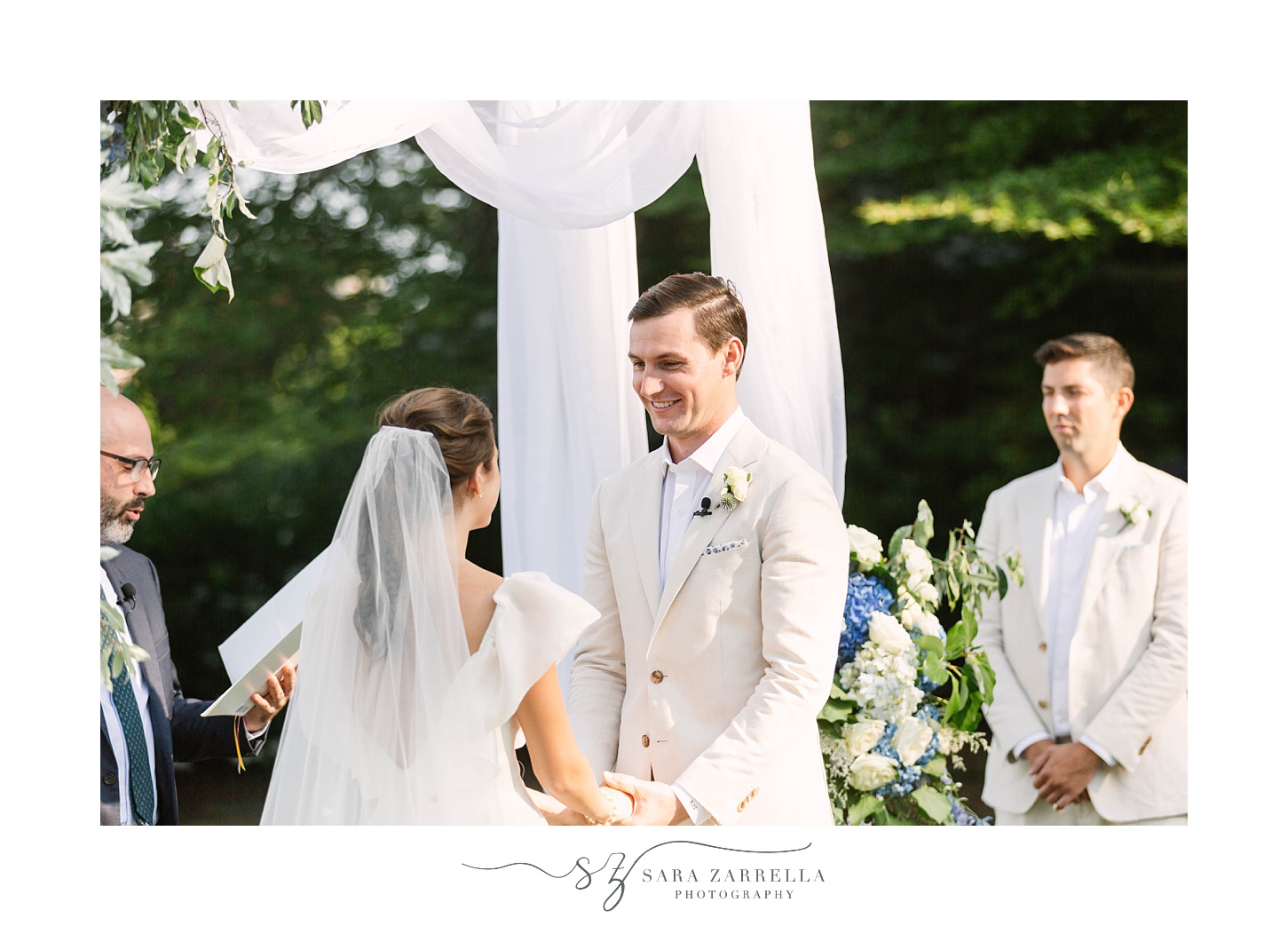 groom smiles at bride holding hands during outdoor ceremony 