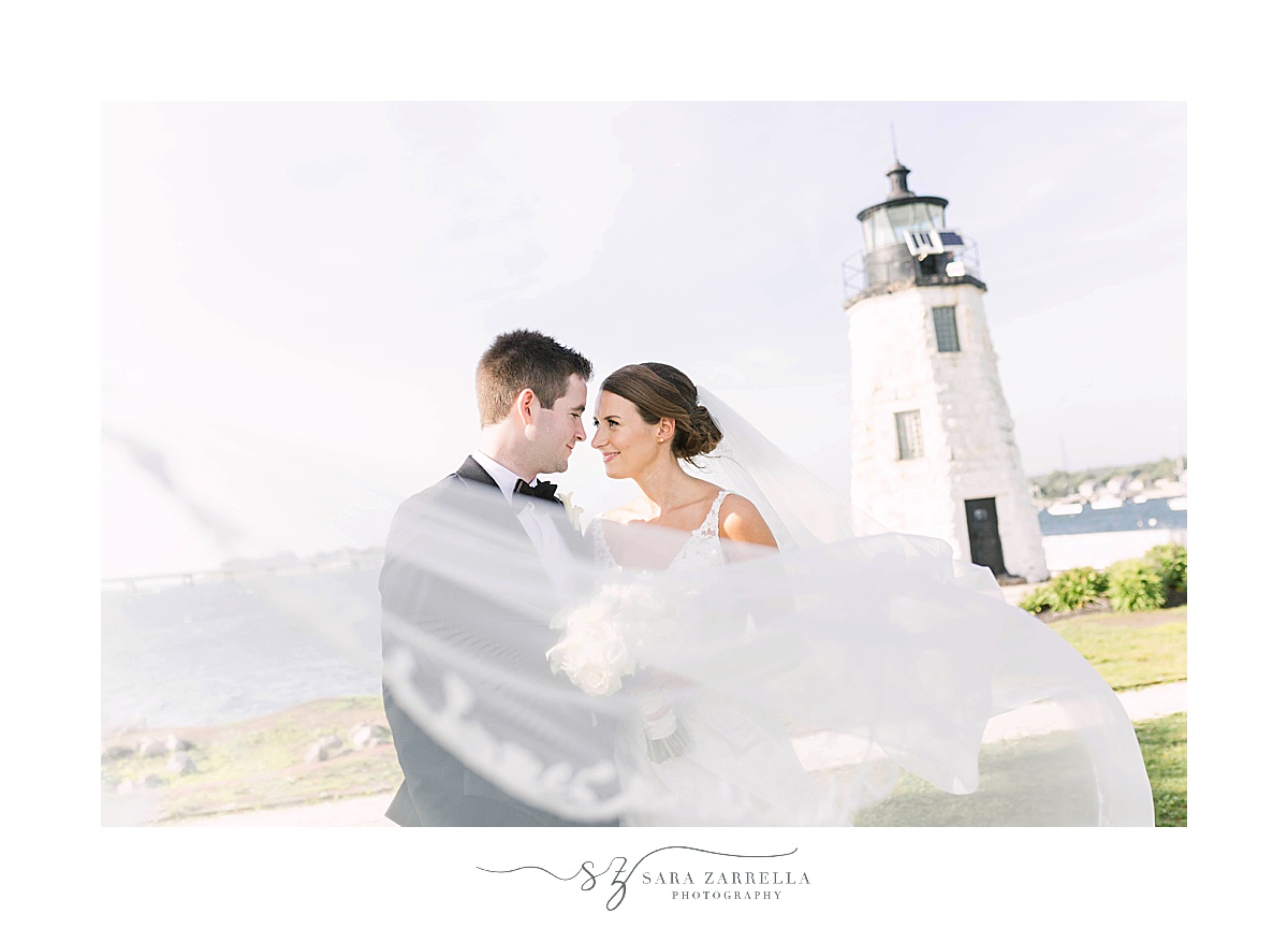 newlyweds pose by lighthouse with bride's veil around shoulders