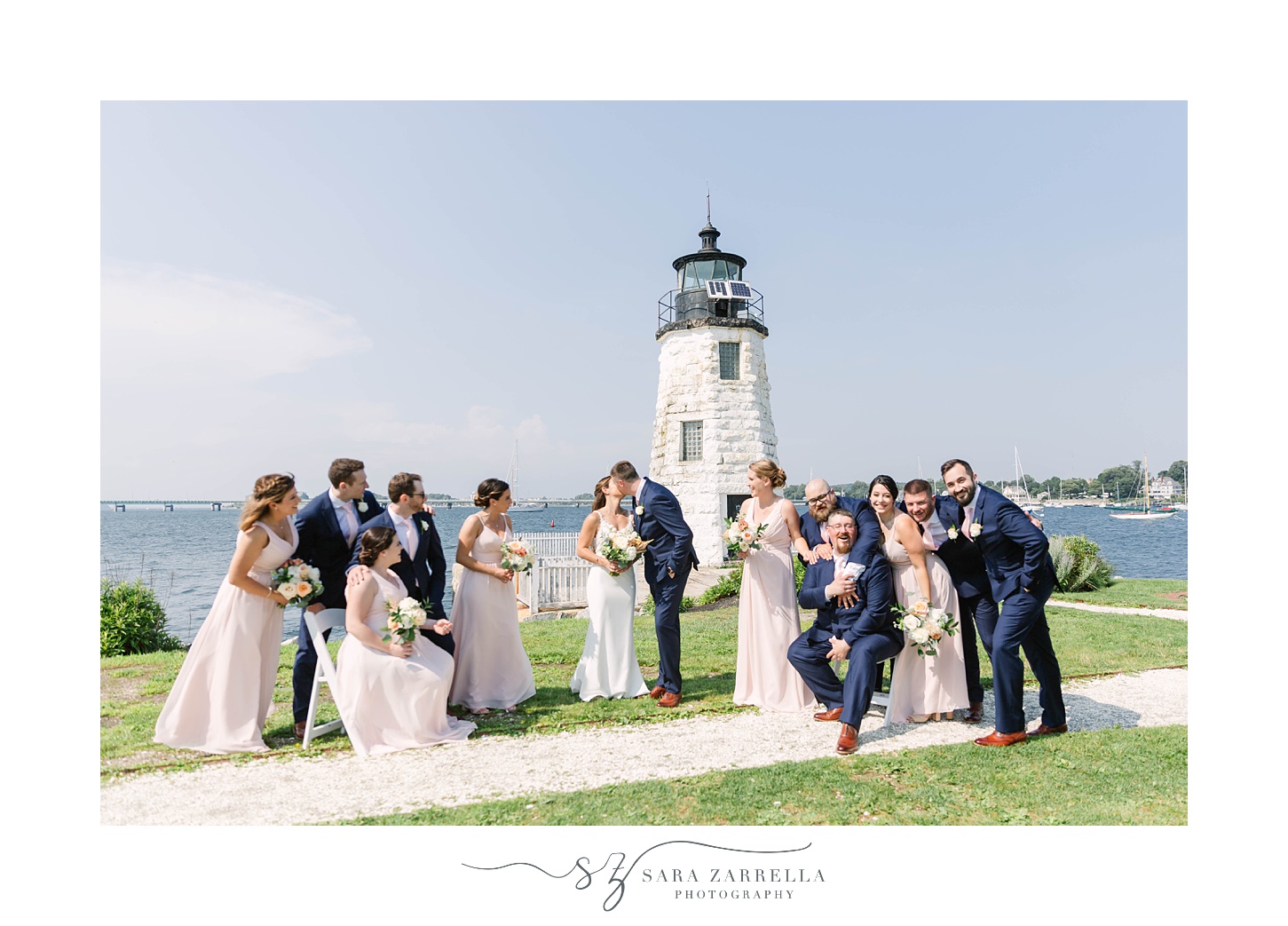 bride and groom kiss while wedding party looks at them by lighthouse in Newport RI