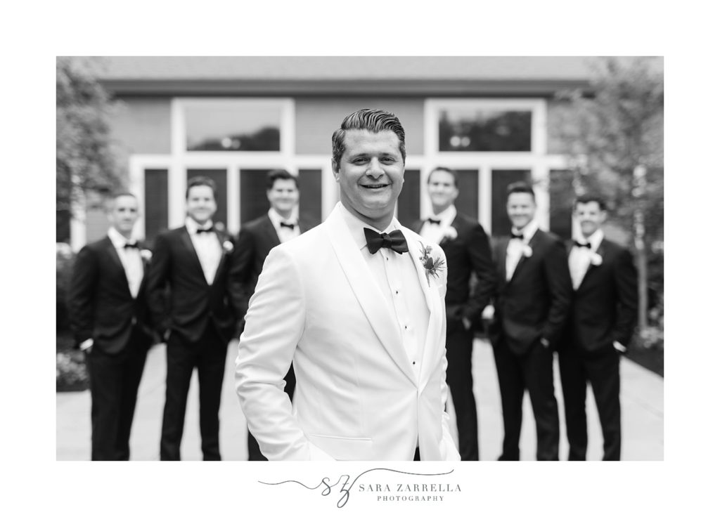 groom poses in white jacket with groomsmen behind him