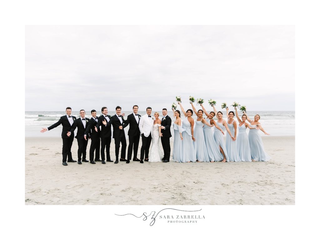 bride and groom pose with wedding party holding bouquets in the air by Narragansett Bay