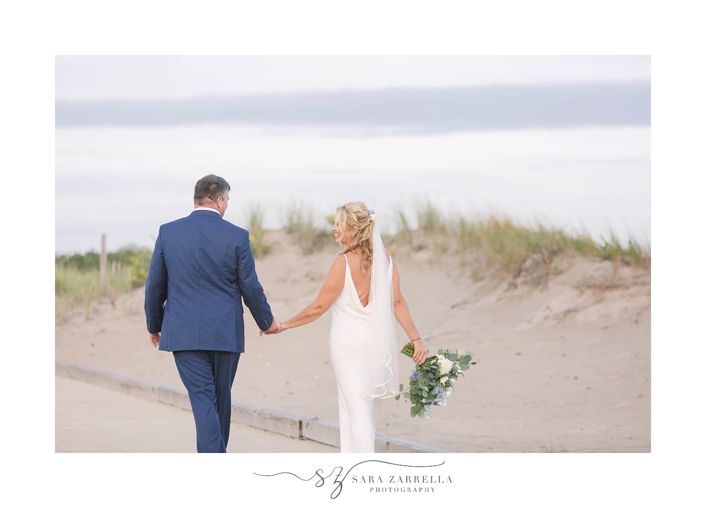 couple holds hands walking through beach dunes in Rhode Island