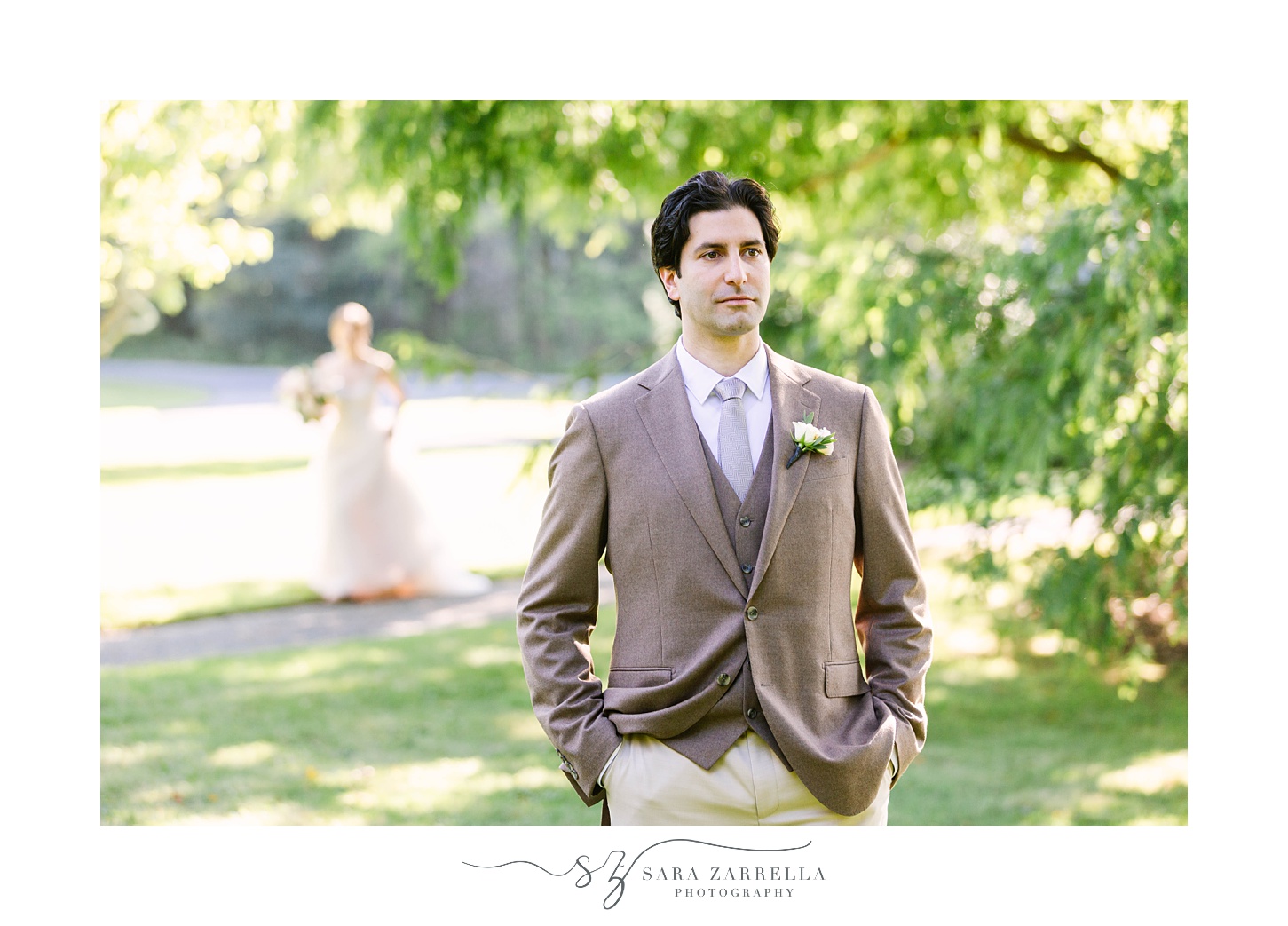groom stands in tan suit with hands in pockets as bride approaches for first look