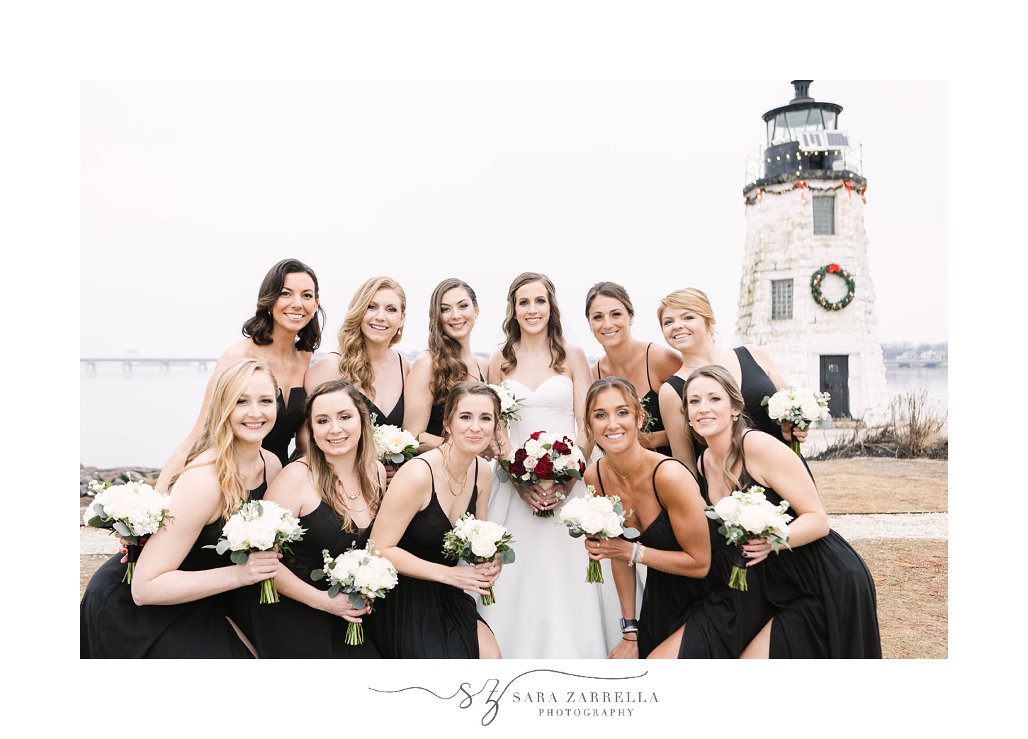 bridesmaids in black dresses hug around bride during photos by lighthouse
