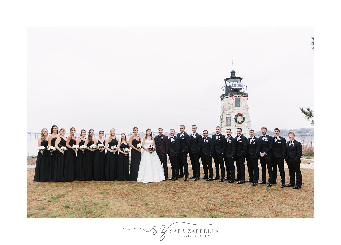 bride and groom stand with bridal party in all-black by lighthouse