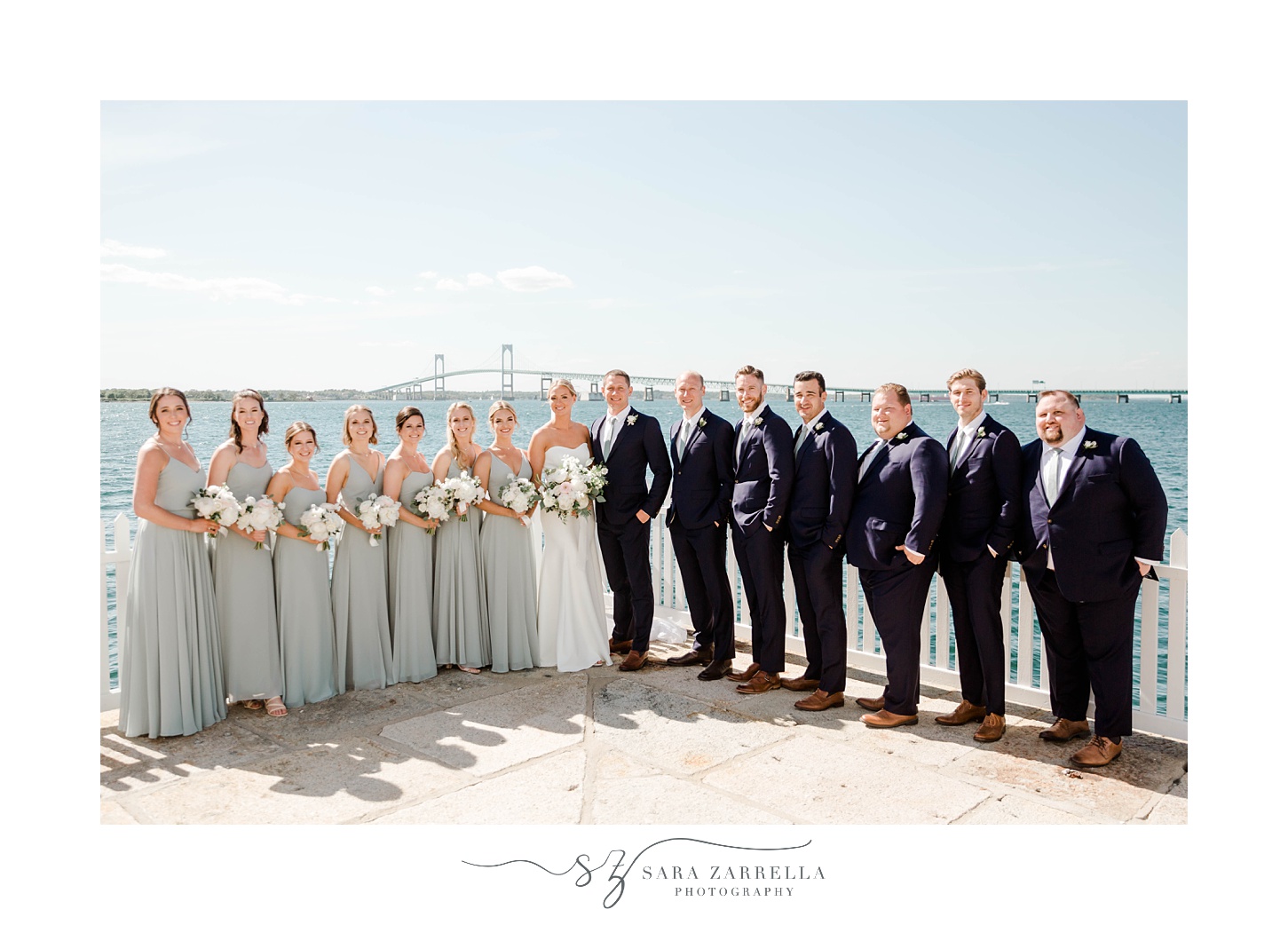 bride and groom lean together with bridal party around them in front of Newport waterfront