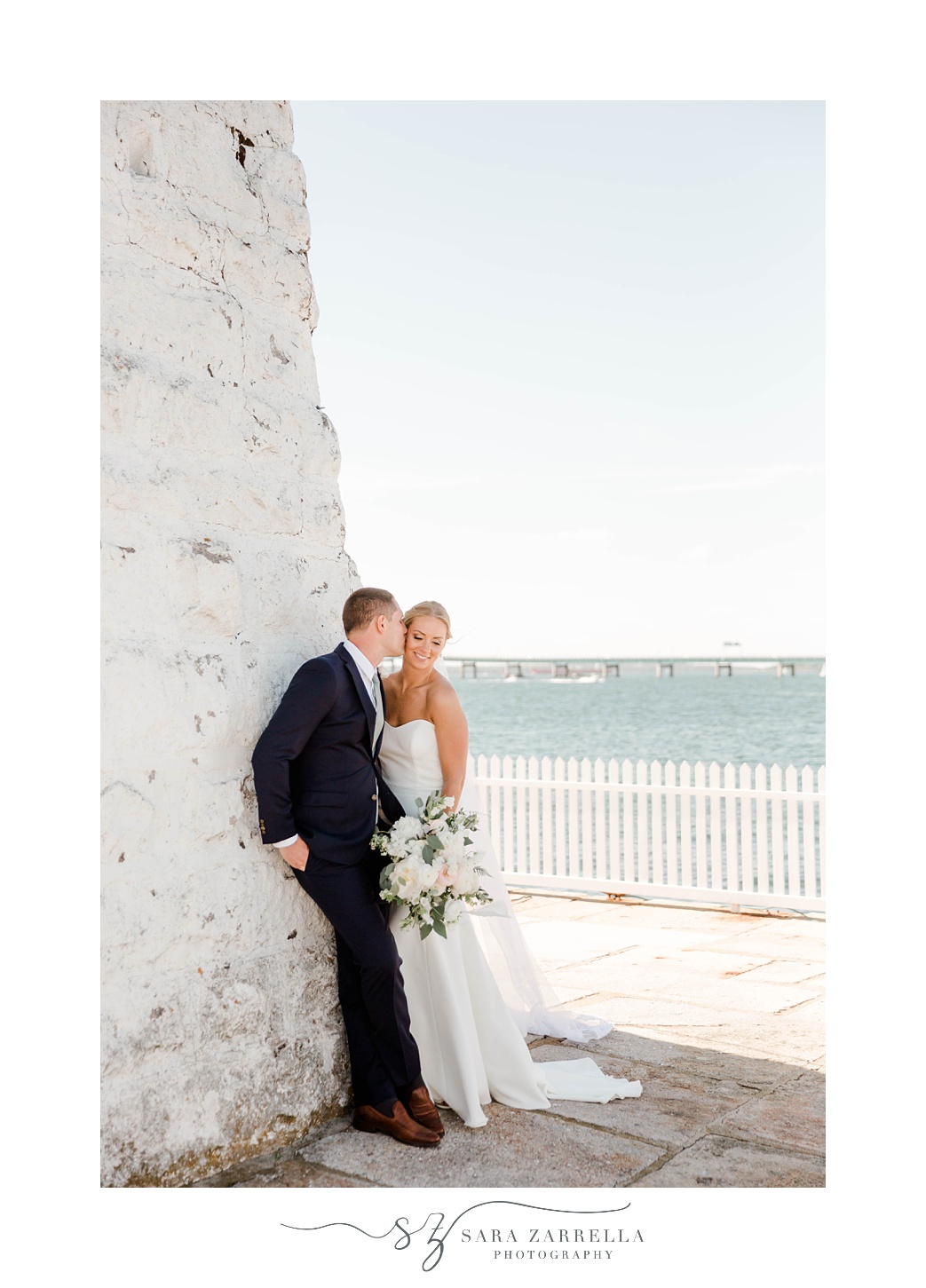 groom leans to kiss bride's cheek against side of white lighthouse in Newport RI