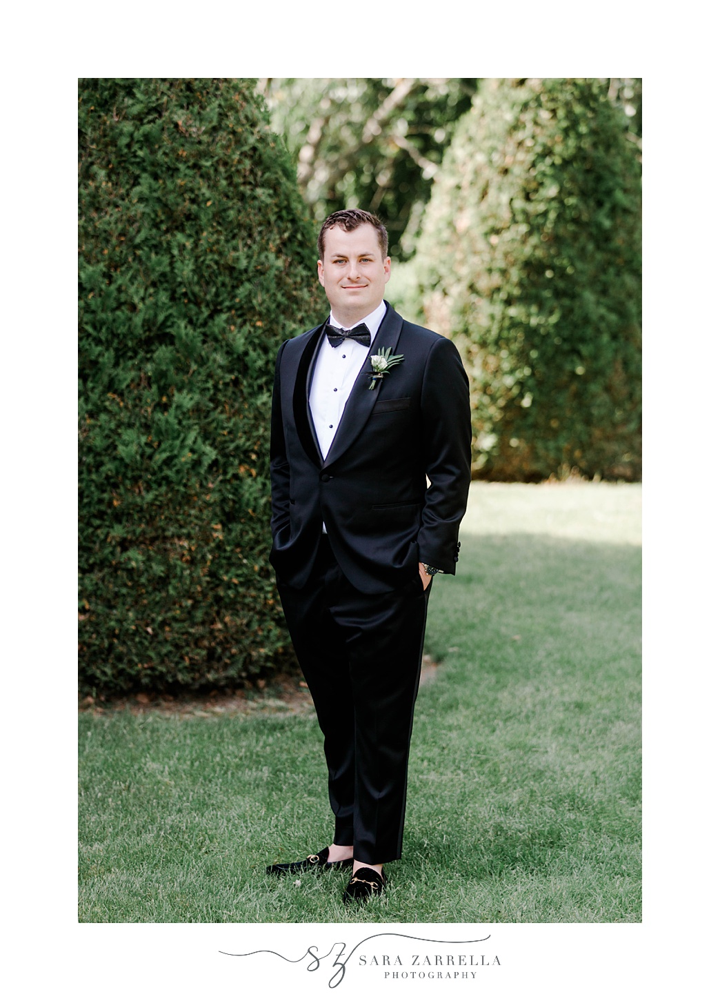 groom in black suit stands with hands in pockets