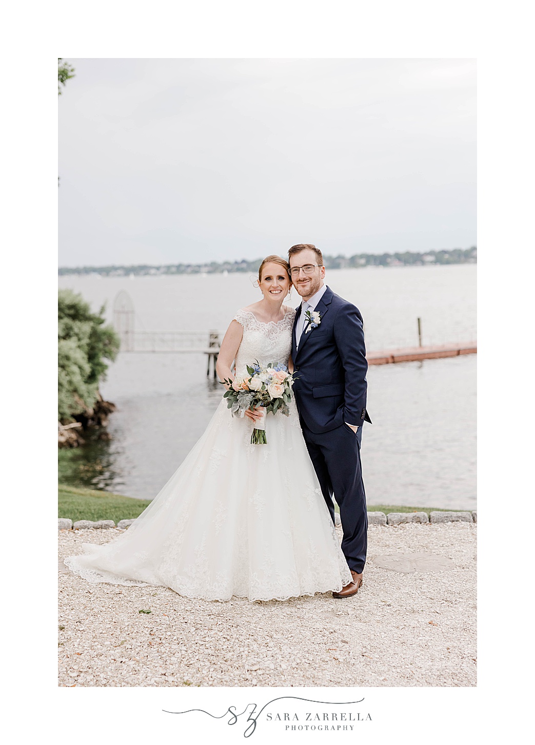 bride and groom stand together in front of water