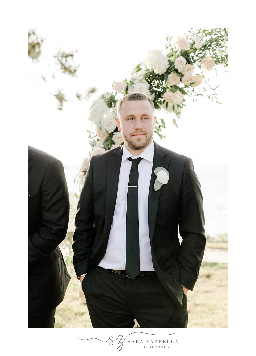 groom stands with hands in pocket of black suit at ceremony 