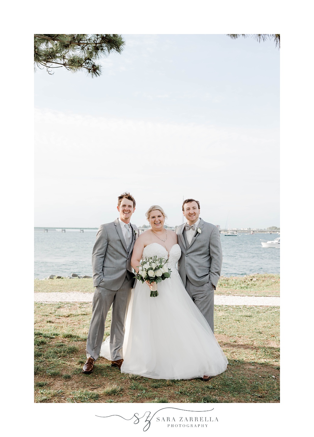 bride poses with groomsmen in grey suits by waterfront 