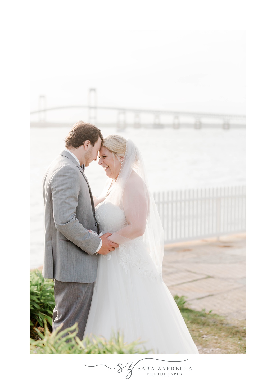 bride and groom lean heads together holding hands with Newport Bridge behind them