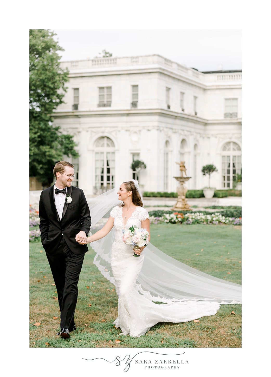 couple holds hands walking on lawn at Rosecliff Mansion with bride's veil trailing behind them