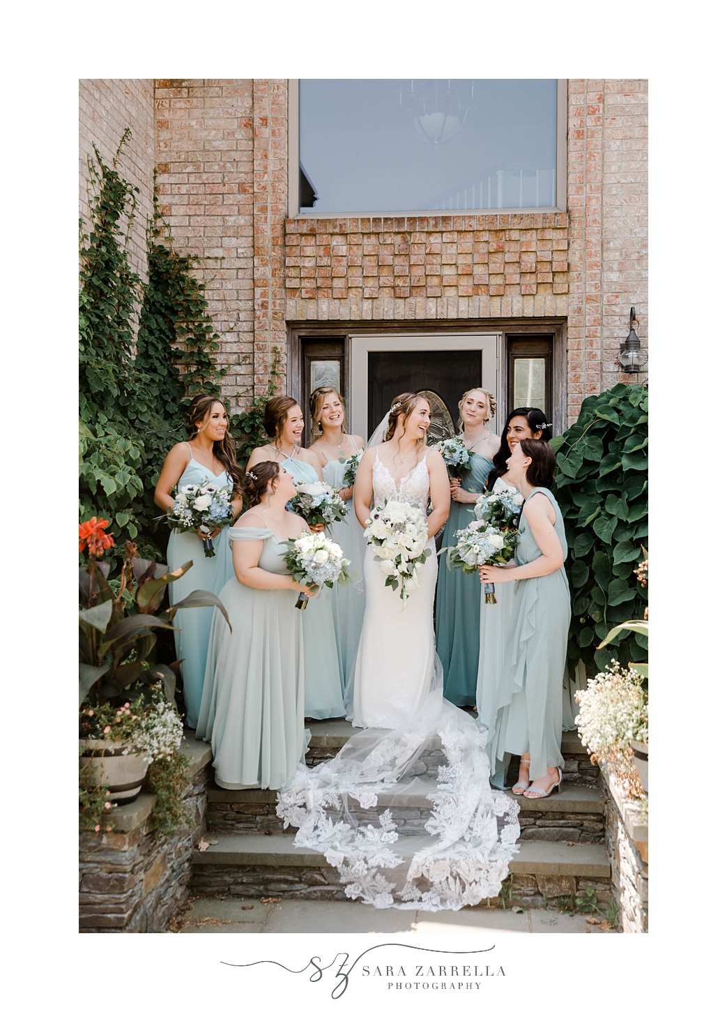 bride stands on steps of brick building with bridesmaids around her in green gowns