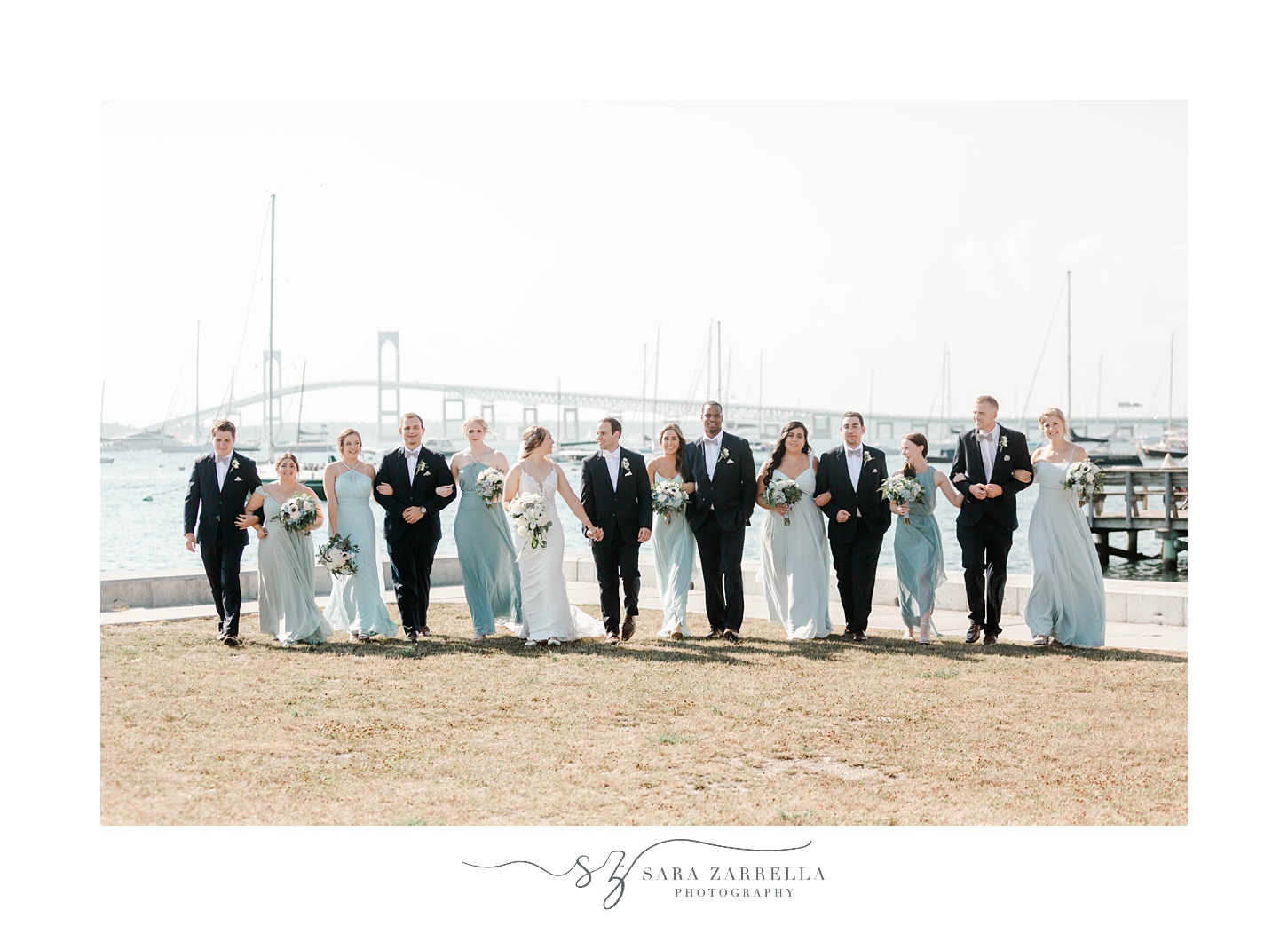 bride and groom walk with wedding party with Newport Bridge behind them at The Bohlin