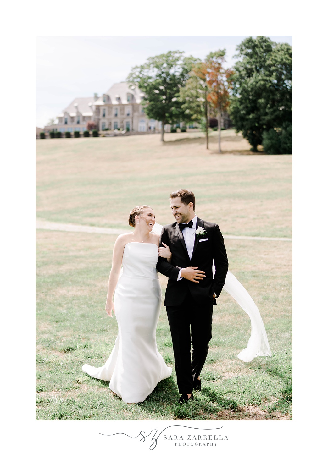 bride smiles over at groom walking on his arm on lawn at Aldrich Mansion