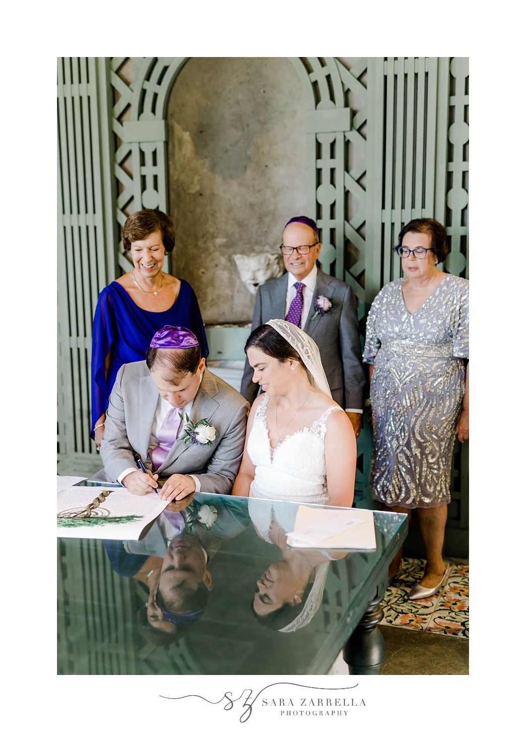 newlyweds sit at table to sign Ketubah with parents behind them at Blithewold Mansion