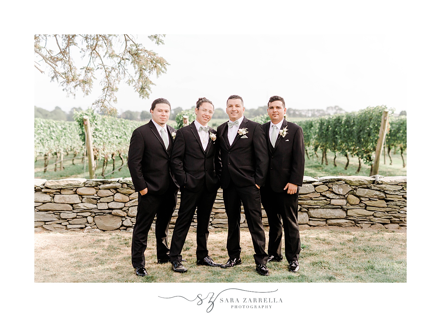 groom and groomsmen in classic black suits stand by stone wall