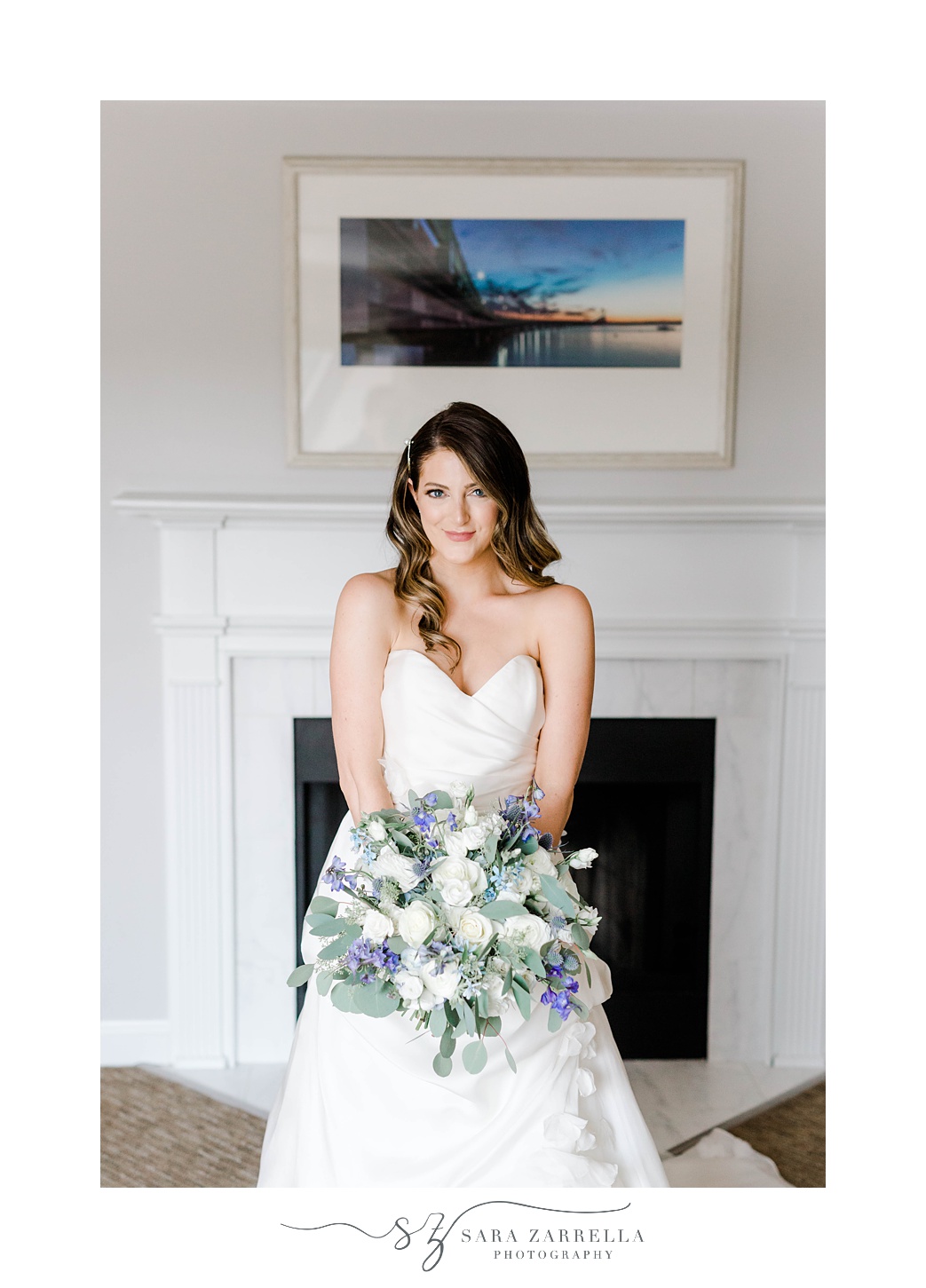 bride stands in front of fireplace holding bouquet of purple and white flowers