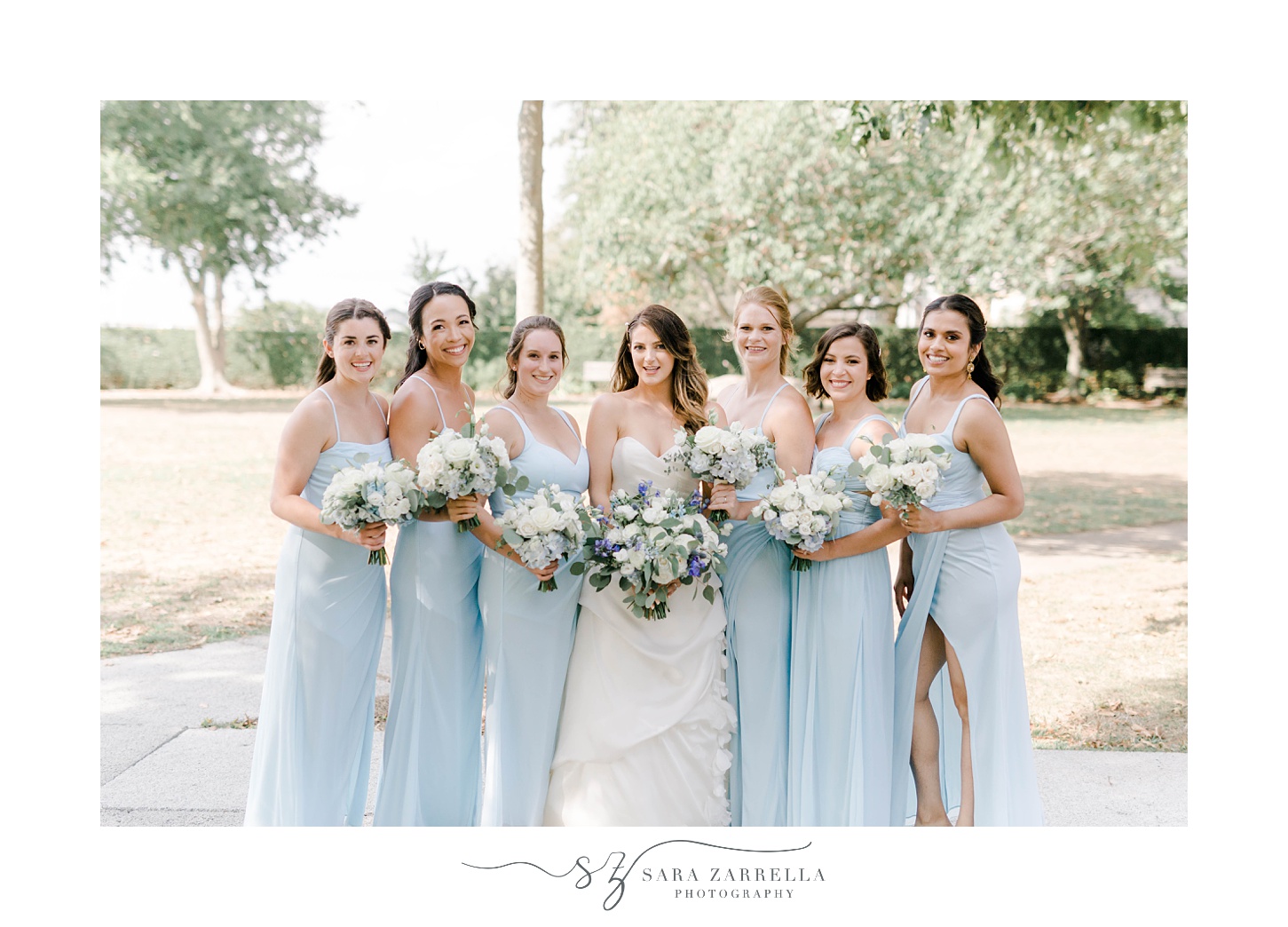 bride poses with bridesmaids in pale blue gowns in Newport RI park