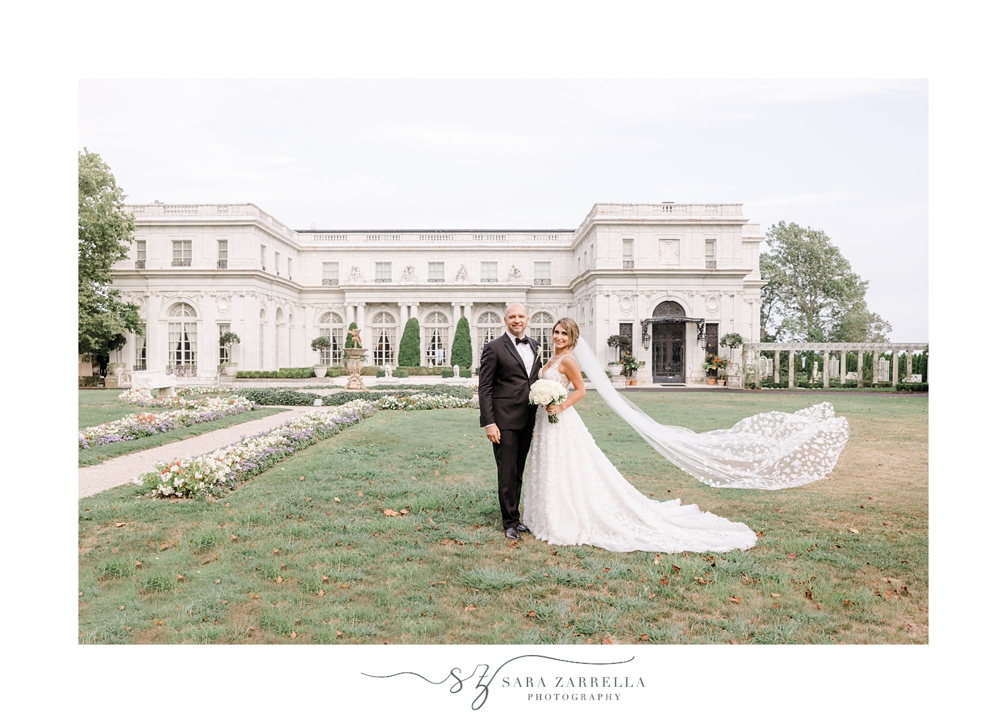 bride and groom stand on lawn at Rosecliff Mansion with bride's veil floating