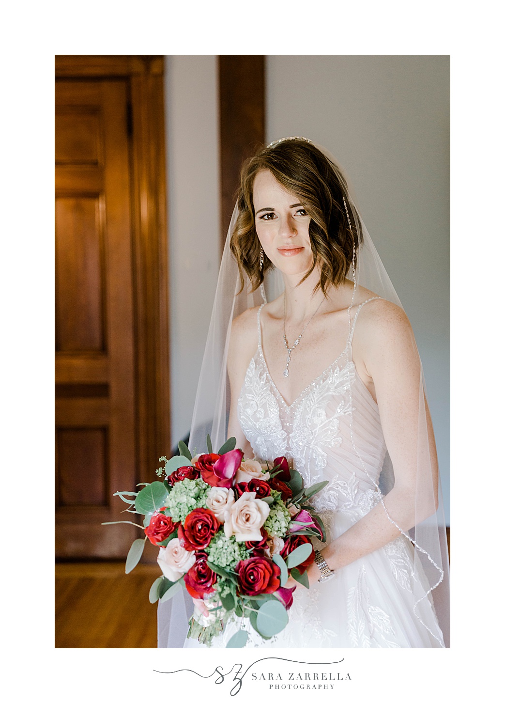 bride stands holding bouquet of pink and red flowers in Eustis Estate library