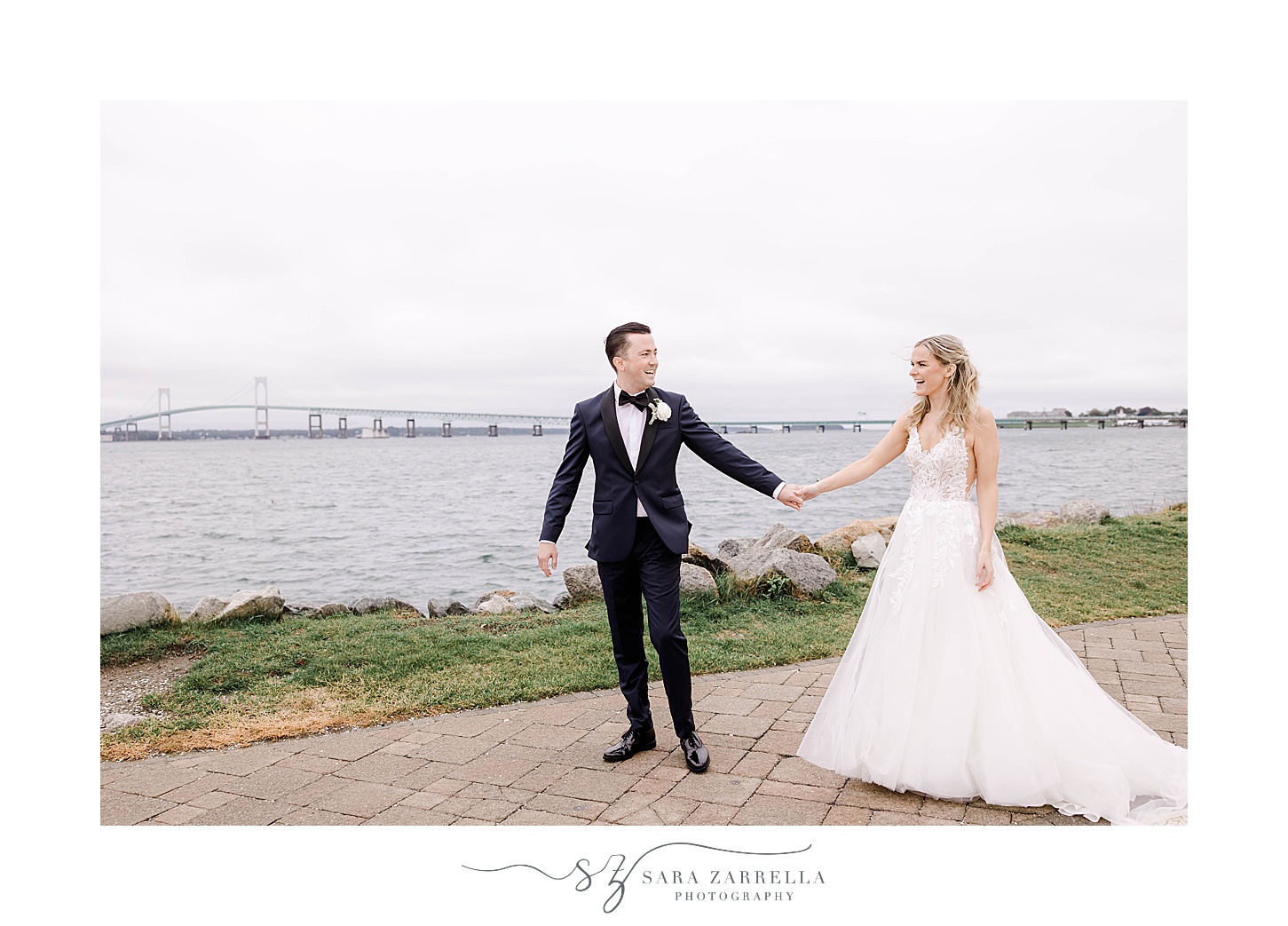 bride and groom hold hands walking with Newport Bridge in background