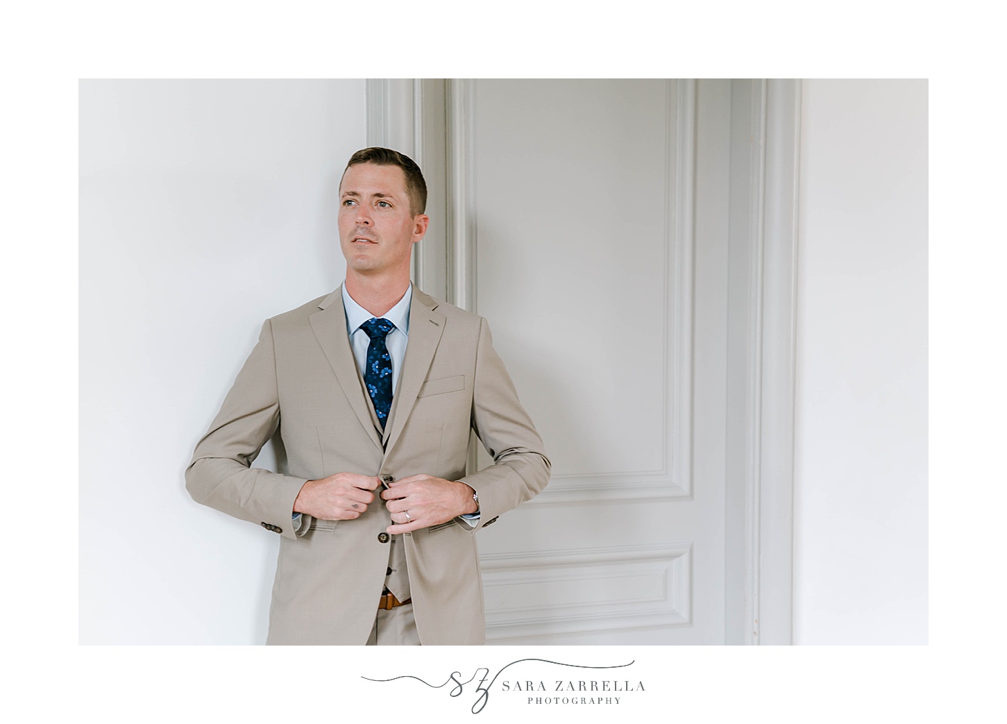 groom stands in tan suit against white wall before Rosecliff Mansion wedding