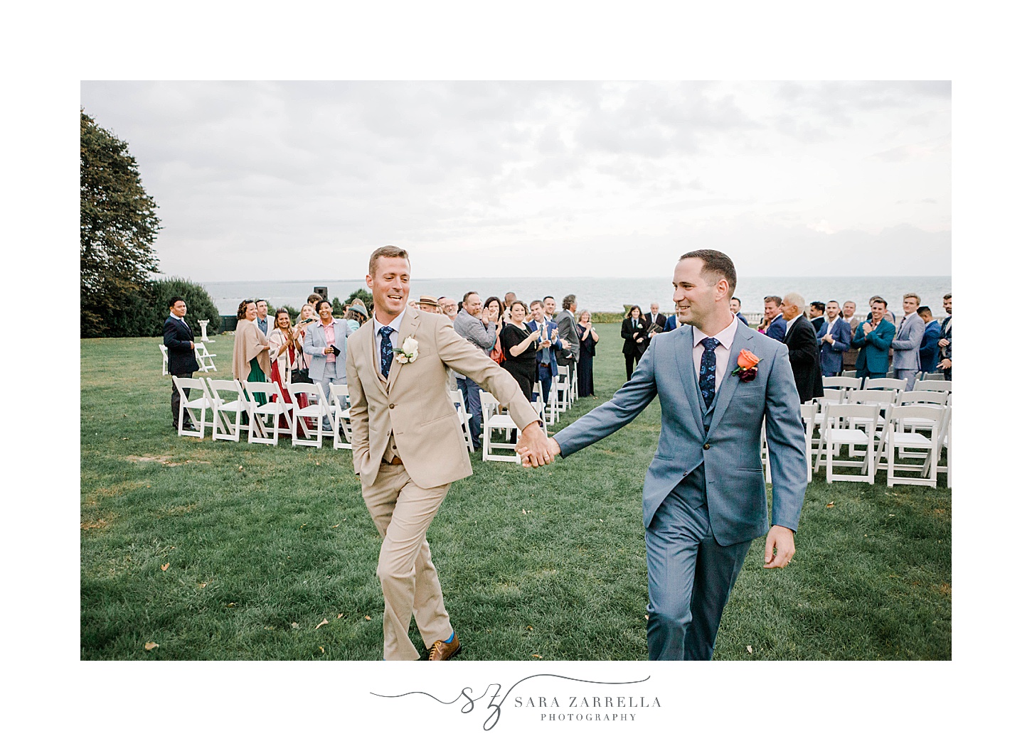 two grooms hold hands walking up aisle at Rosecliff Mansion
