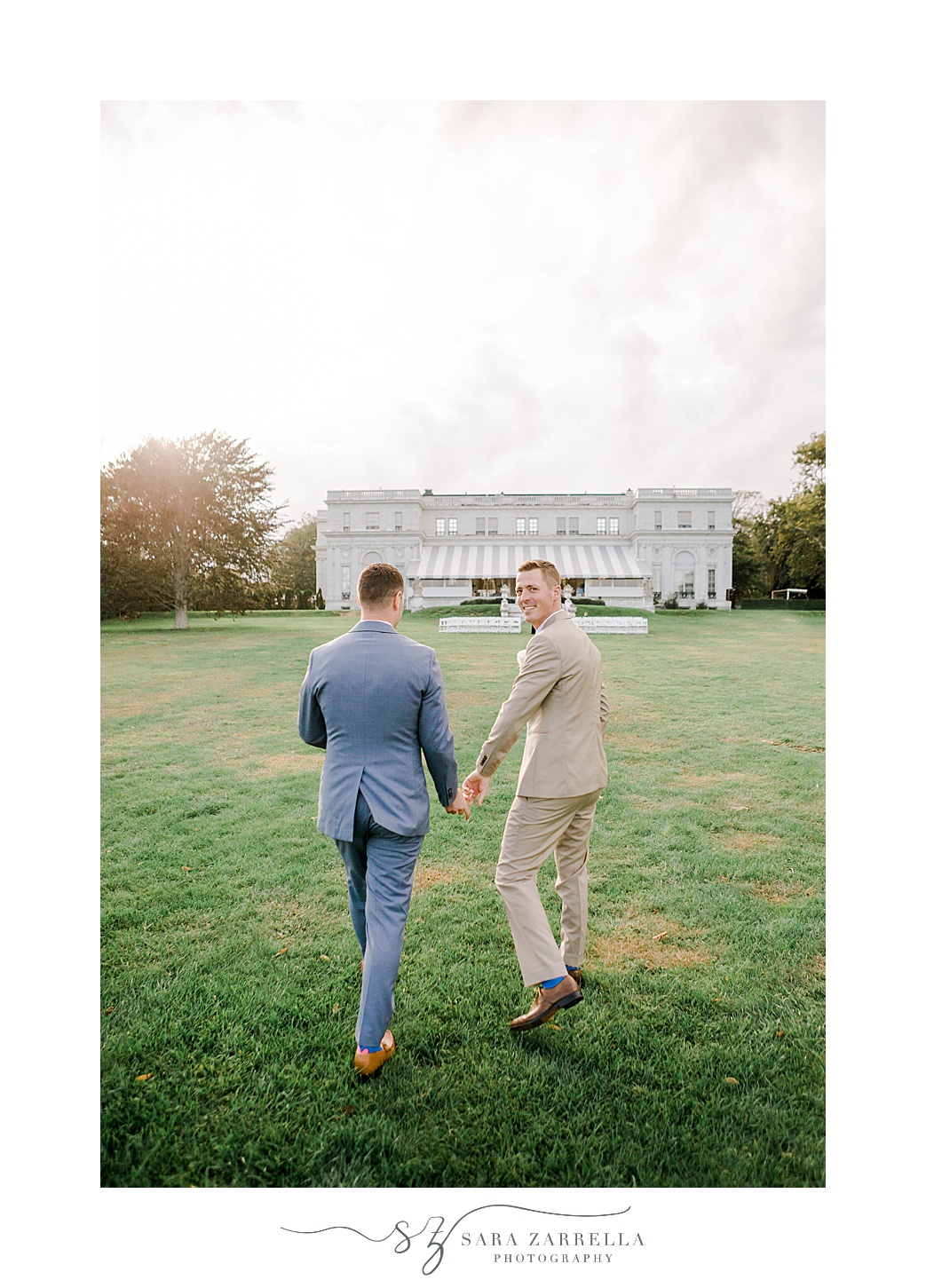 grooms hold hands walking across lawn looking back at photographer