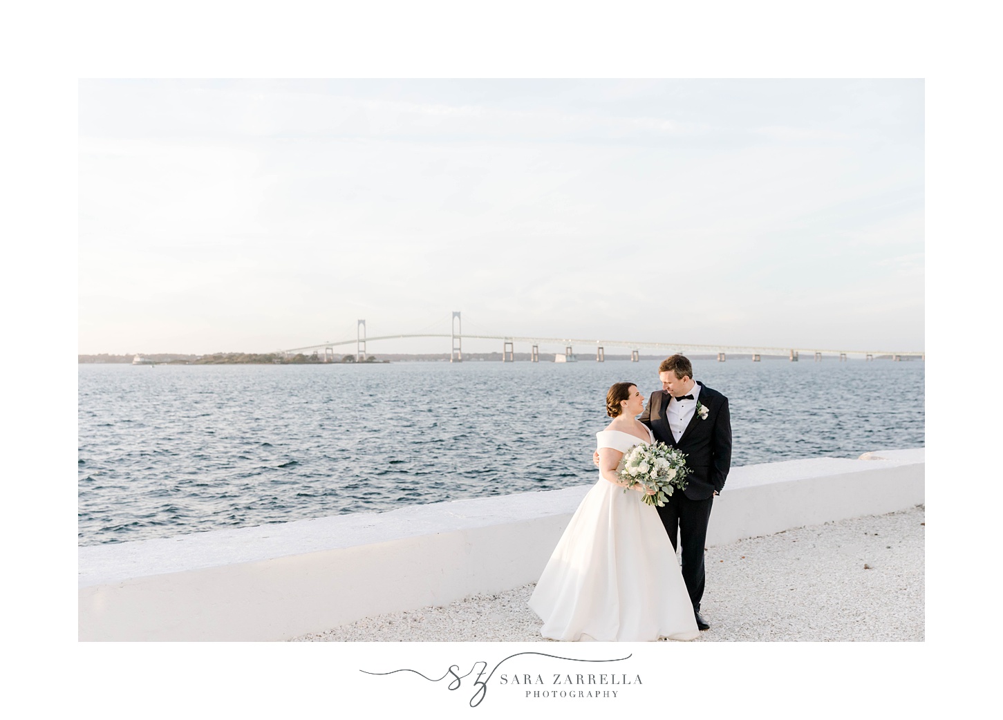 bride and groom hug along white wall with Narragansett Bay behind them