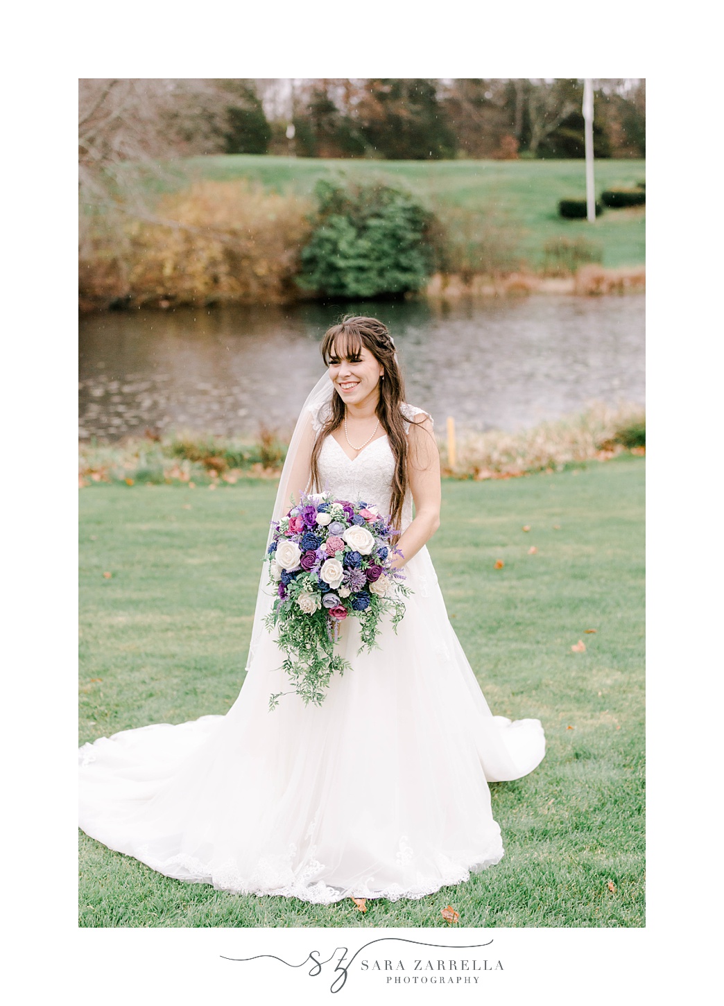 bride stands in front of pond holding bouquet of purple flowers 