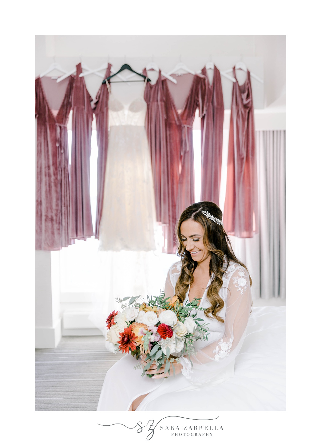 bride sits in front of hanging dress in window looking down at bouquet