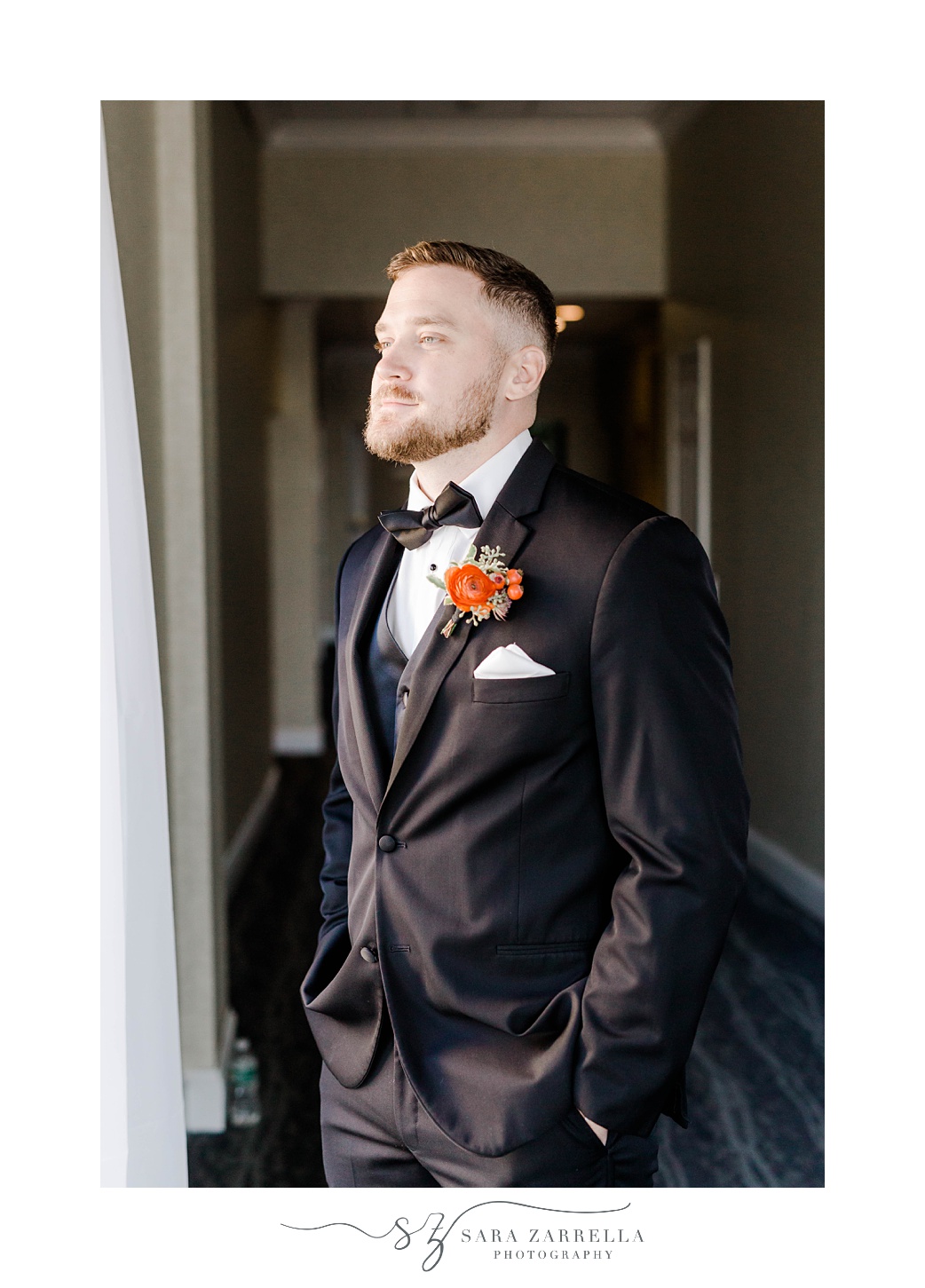 groom stands looking out window at Newport Harbor Island Resort with hands in pocket of suit