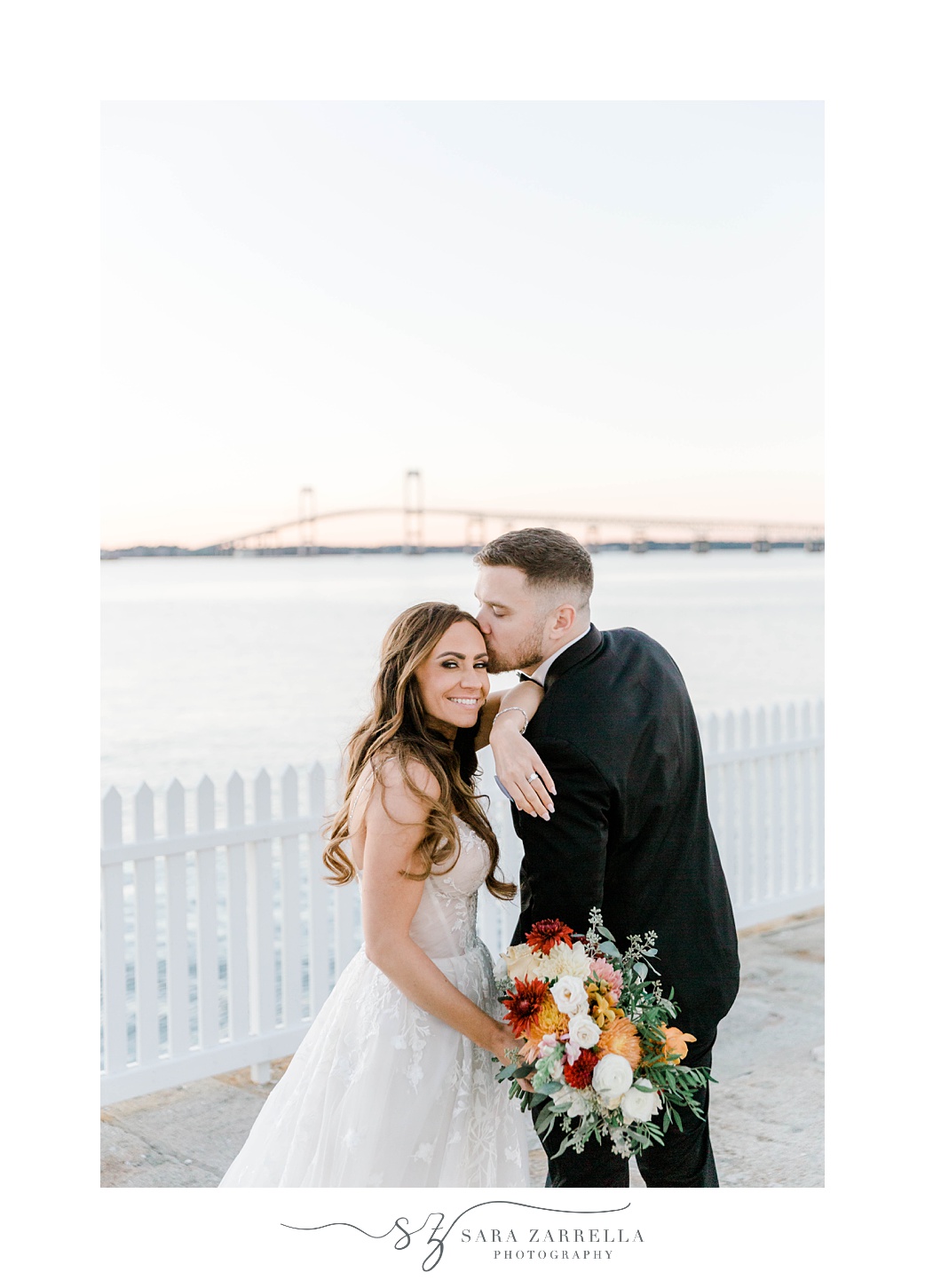 groom leans to kiss bride's forehead with Newport Bridge behind them 