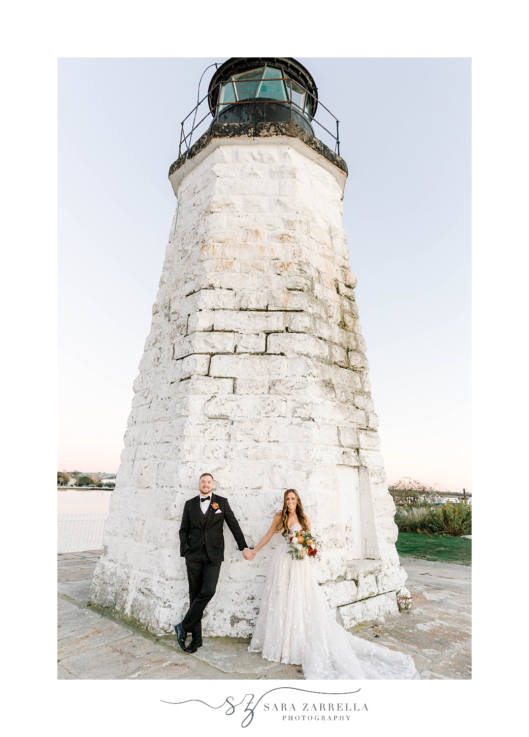 newlyweds hold hands leaning against Newport Lighthouse 