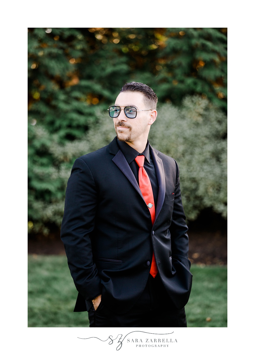 groom stands with hands in pockets of black suit with red tie and Tony Stark glasses 
