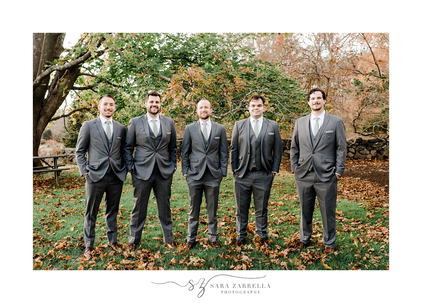 groom stands with groomsmen on fallen leaves with hands in pockets of suits 