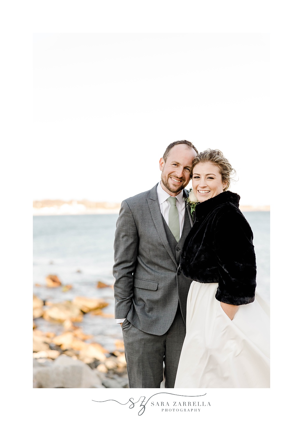 groom hugs bride in black fur on beach in Rhode Island 