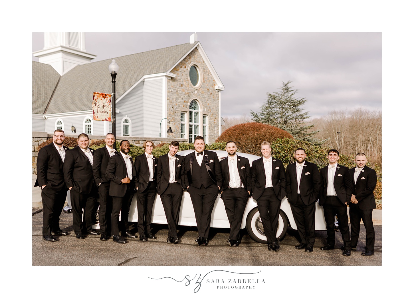 groom and groomsmen in black suits lean against custom white car outside RI church 