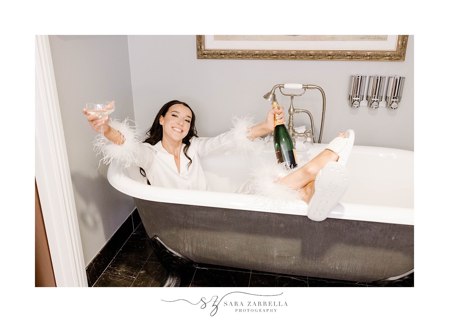 bride sits in bathtub in white robe holding champagne 
