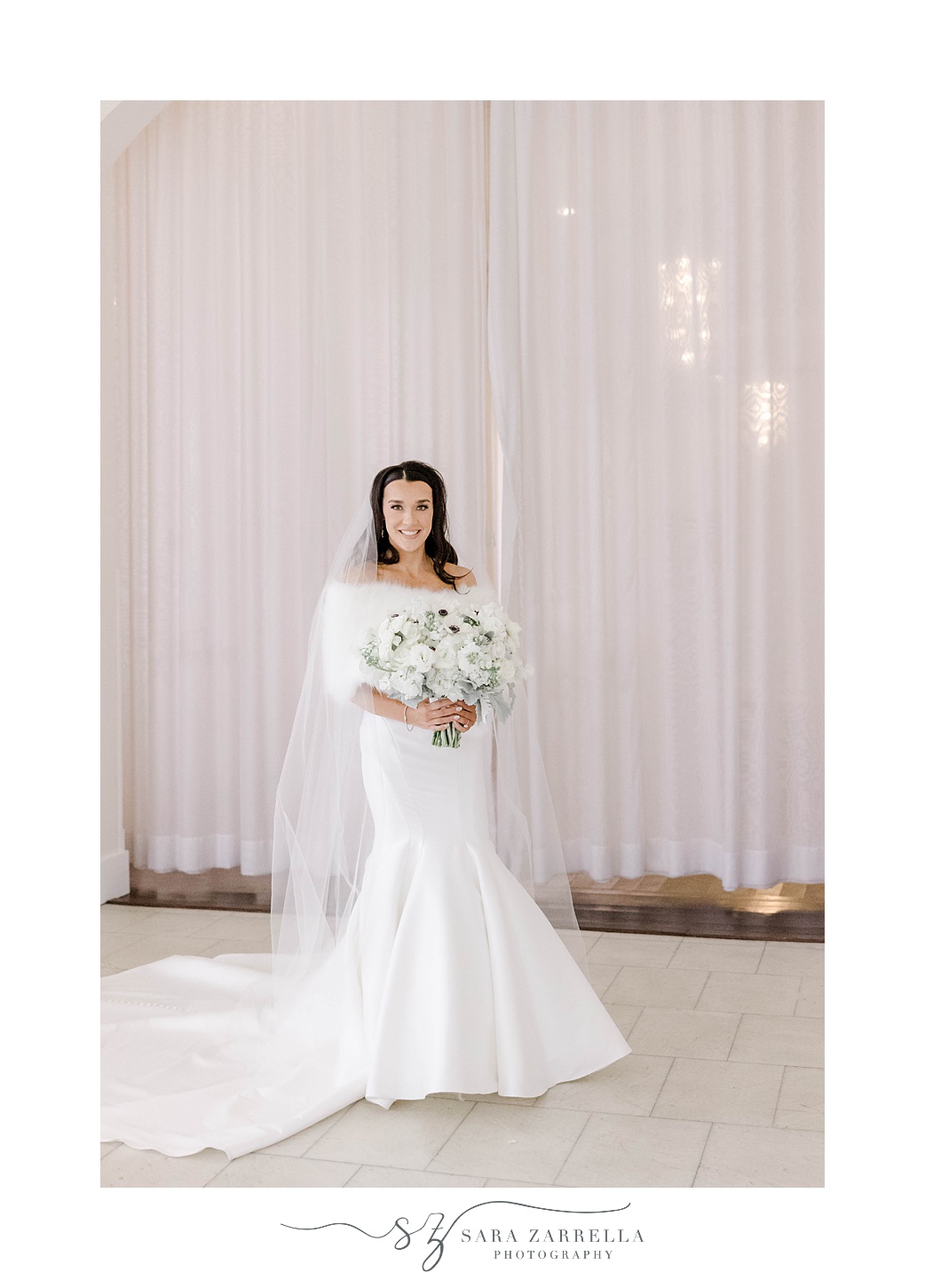 winter bride poses in fitted gown with white fur and bouquet of white anemones