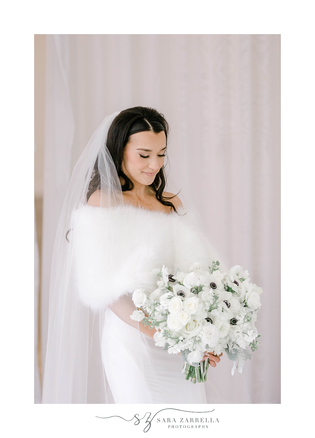 winter bride looks down at bouquet of white anemones with veil around shoulders 